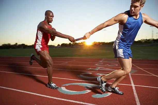 Two athletes in a relay race passing the baton on a running track at sunset, symbolizing teamwork and seamless collaboration.
